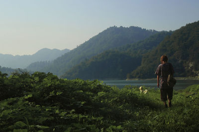 Rear view of woman standing on grassy field by lake against mountains