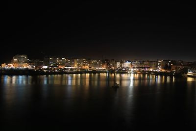 Illuminated buildings by sea against sky at night