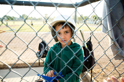 Portrait of boy wearing baseball helmet while sitting in dugout seen through chainlink fence