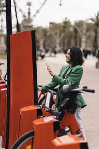 Side view of cheerful female with smartphone using public red terminal while paying for bike sharing on street of city