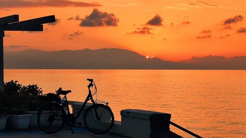 Silhouette people riding bicycle by sea against orange sky