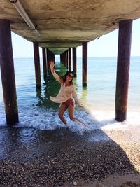 Full length of playful woman dancing below pier at beach