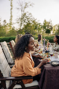 Side view of happy woman laughing while having food with friends at dining table in back yard