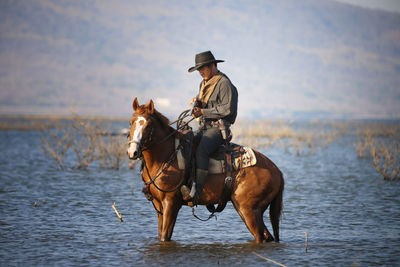 Side view of cowboy riding horse at in lake against mountains