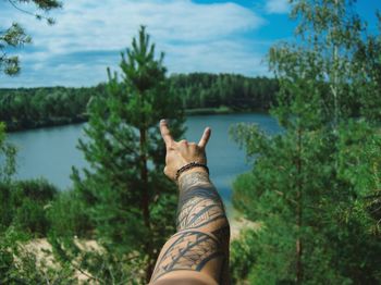 Cropped hand of man with tattoo gesturing against lake
