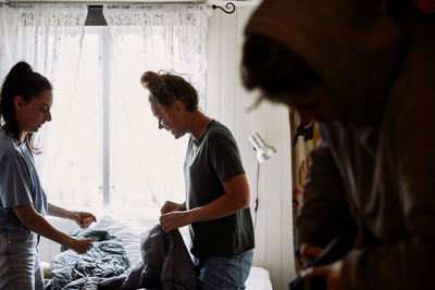 Side view of female friends folding blankets while kneeling on bed against window in cottage