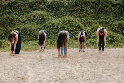 Women with yoga instructor practicing uttanasana on sand against plants