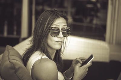 Portrait of mature woman wearing sunglasses using mobile phone while sitting on sofa