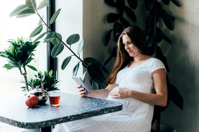 A pregnant girl is sitting at a table in a cafe with a mobile gadget and reading.