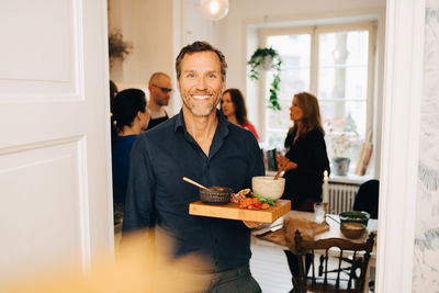 Portrait of smiling man with serving tray standing against friends in party at home