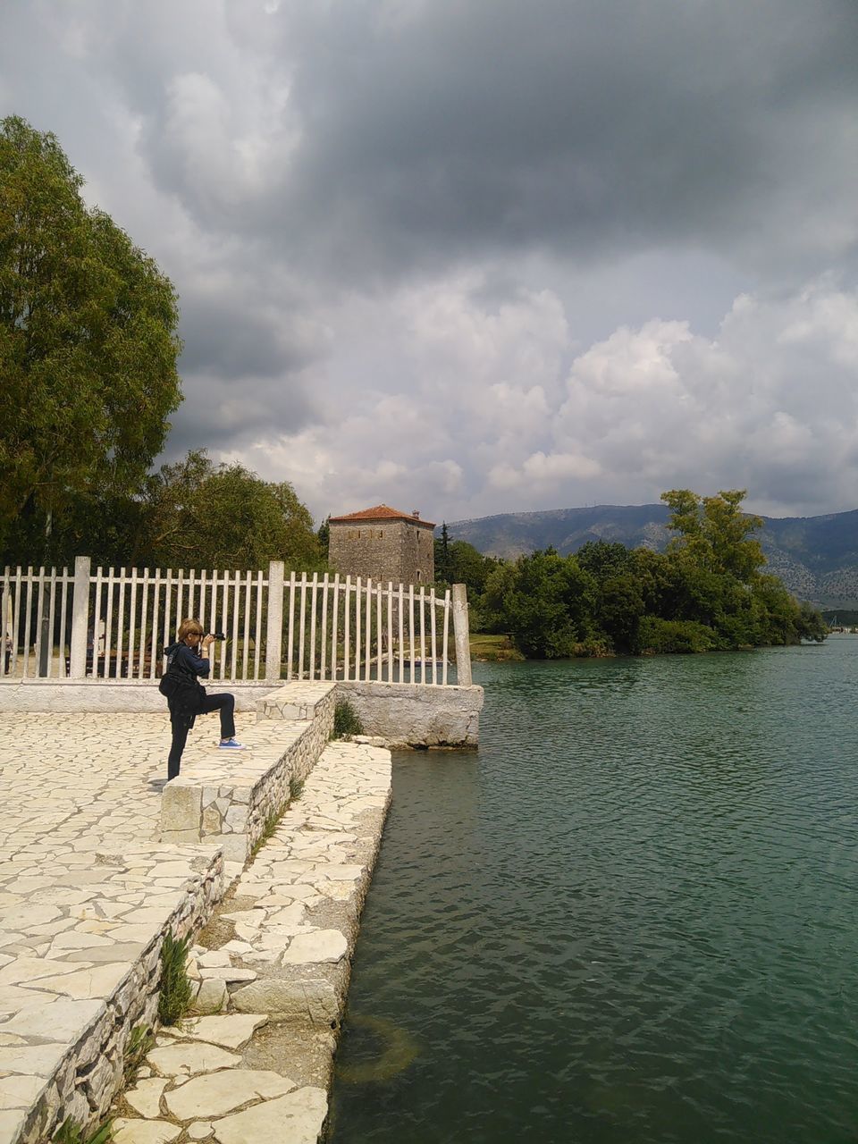 REAR VIEW OF MAN STANDING ON RIVERBANK AGAINST SKY