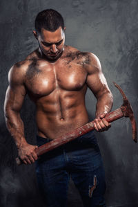 Shirtless muscular worker holding pick axe while standing against wall