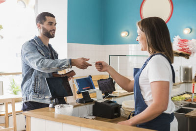 Young female owner giving credit card to male customer while standing at checkout in restaurant