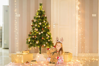 Cute smiling girl sitting by illuminated christmas tree