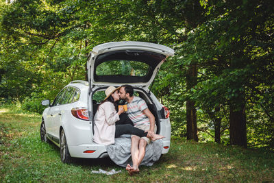 Young couple sitting in boot of car in forest, drinking coffee.