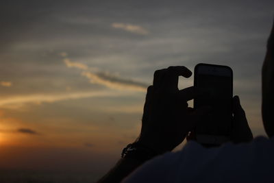 Cropped hands photographing against sky at sunset