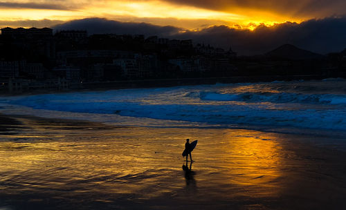 Silhouette person with surfboard standing at beach against sky during sunset