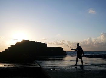 Man with fishing rod at pier against sky during sunset