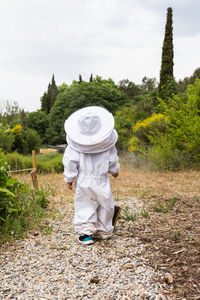 Back view of unrecognizable kid in white beekeeper protective costume standing near metal bee smoker in village