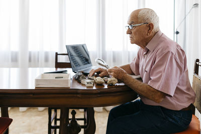 Retired senior man using magnifying glass for research of fossil and mineral at home