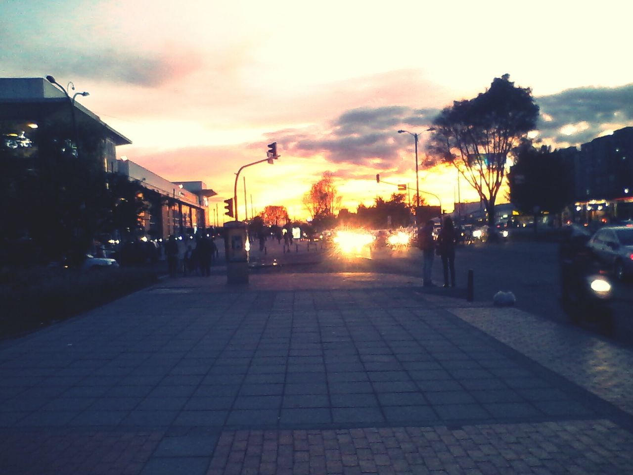 sunset, building exterior, sky, architecture, built structure, street, city, street light, cloud - sky, tree, silhouette, transportation, incidental people, city life, city street, orange color, road, car, sunlight, cobblestone