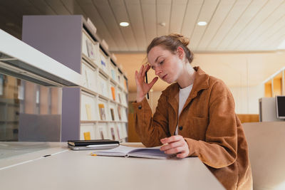 Young woman in casual clothes with hair bun touching head and writing in planner while sitting at table and studying in university library