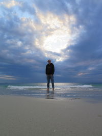 Full length of man standing on shore at beach against cloudy sky during sunset