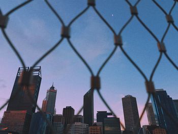 Low angle view of modern buildings against sky seen through chainlink fence