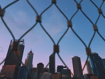 Low angle view of modern buildings against sky seen through chainlink fence