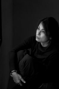 Woman looking away while crouching on chair against black background