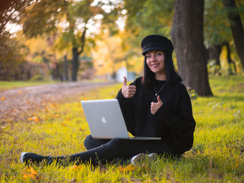 Portrait of young woman gesturing thumbs up while using laptop at public park during autumn