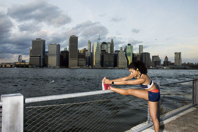 Female athlete exercising by river against sky in city during sunset