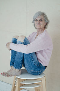 Smiling senior woman with gray hair sitting on chairs in front of wall
