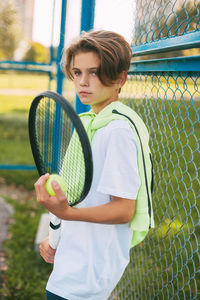 Boy with tennis racket standing by fence