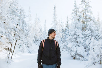 Young male tourist in outerwear with backpack looking away against snowy spruces on sunny winter day in valley of the ghosts in monts valin national park in quebec, canada