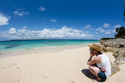 Side view of woman crouching at beach against blue sky during sunny day