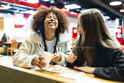 Cheerful woman holding disposable cup with friend in cafe