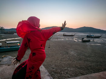 Rear view of girl gesturing while standing at beach against sky during sunset