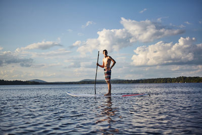 Full length of shirtless man paddleboarding in sea against sky