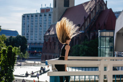 Side view of playful young woman tossing hair on balcony against buildings in city