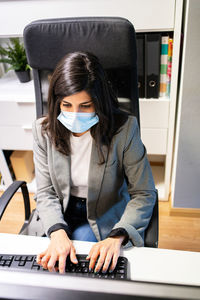 From above confident young female specialist in formal suit and protective mask sitting at desk and using computer while working in modern workspace