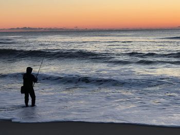 Silhouette man fishing on beach against sky during sunset