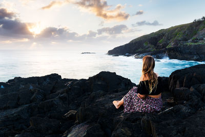 Rear view of woman sitting on rock by sea against sky