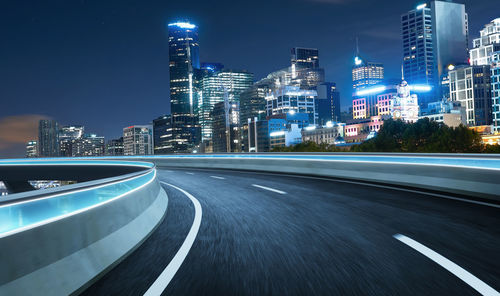 Illuminated road by buildings against sky at night