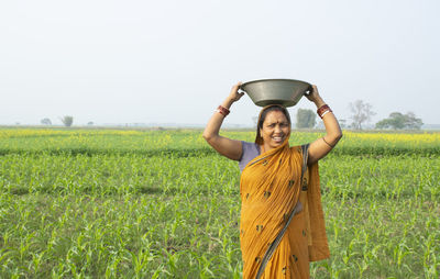 Portrait of smiling young woman farmer standing on field holding fertilizer basket on her head