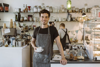 Smiling male owner wearing apron holding coffee cup at cafe