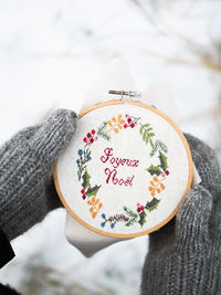 Close-up of hands in gray knitted mittens hold a hoop with christmas embroidery joyeux noel