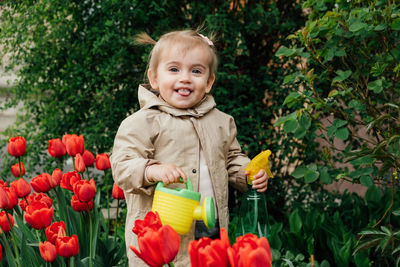 Spring gardening. cute toddler little girl in raincoat watering red tulips flowers in the spring