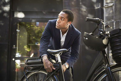 Young male commuter locking electric bicycle while looking away against building in city