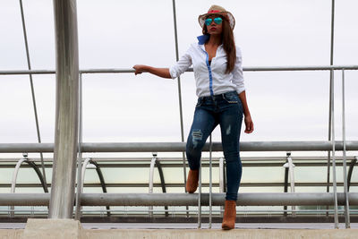 Portrait of young woman standing by railing against sky